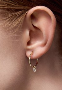 Złota kolczyk w kształcie obręczy z przezroczystym, sercowym zawieszeniem, mający polerowane wykończenie i gładką teksturę, noszony na płatku ucha.