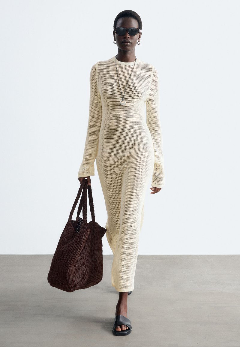 Femme portant une longue robe en maille crème, des lunettes de soleil noires, des sandales noires à enfiler, portant un grand sac cabas tricoté marron foncé.
