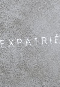 Grijze textuur achtergrond met reliëfletters die "EXPATRIÉ" spellen. De letters zijn wit en glanzend, wat contrasteert met het matte oppervlak.