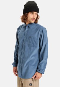 Blauwe corduroy button-up shirt met een borstzak, lange mouwen en een relaxte pasvorm, gecombineerd met een zwarte gebreide beanie.
