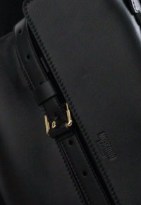 Sac en cuir noir à texture lisse, avec une boucle en métal doré et un logo embossé. Design minimaliste avec une forme structurée.