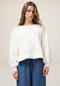 Felpa bianca con maniche a palloncino, caratterizzata da dettagli in pizzo floreale sui polsini. Abbinata a pantaloni in denim a gamba larga.