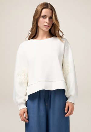 Felpa bianca con maniche a palloncino, caratterizzata da dettagli in pizzo floreale sui polsini. Abbinata a pantaloni in denim a gamba larga.