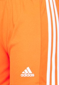 Shorts de sport orange en tissu lisse, avec trois rayures blanches sur le côté et un logo Adidas blanc sur l'ourlet.