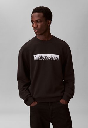 Mladý muž v čiernej mikine Calvin Klein pozerá doľava, ruky v vreckách, stojí pred jednofarebným sivým pozadím.