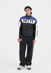 Svart läderjacka med blåa och vita accenter, med siffran "1971" på bröstet och text på ärmen. Mörka jeans och sneakers.