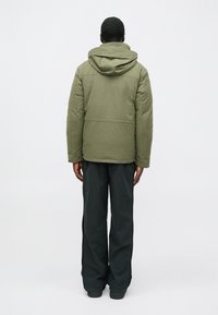 Gepolsterte grüne Jacke mit Kapuze, Rückansicht. Sie hat einen geraden Schnitt mit Nahtdetails und wird mit weiten schwarzen Hosen kombiniert.