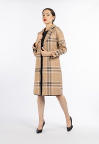 Donna che indossa un lungo cappotto a quadri beige su un vestito nero, in piedi con una gamba tesa e tenendo il colletto del cappotto, su uno sfondo bianco.