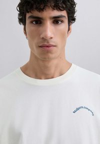 Camiseta blanca de algodón con cuello redondo y texto bordado en azul "Scalpers company" en el pecho. Textura lisa y uniforme.