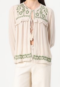 Blusa beige con bordado floral verde, bordes con volantes y un corte en forma de llave. Presenta mangas largas y un ajuste relajado.