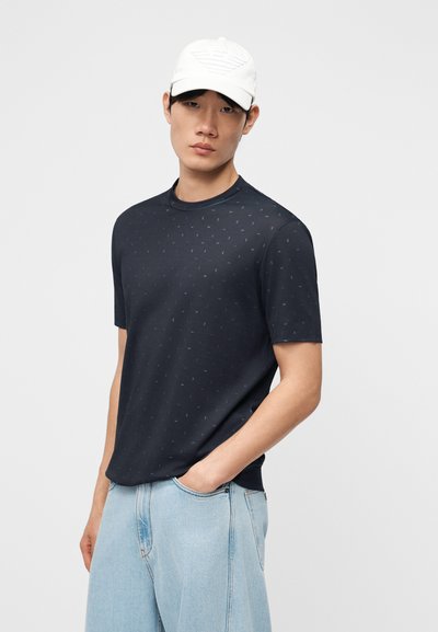 Νέος άνδρας που φοράει λευκό καπέλο, σκουρόχρωμο μπλε κοντομάνικο πουκάμισο με διακριτικές τελείες και ανοιχτόχρωμο μπλε τζιν, στέκεται με το ένα χέρι στην τσέπη.