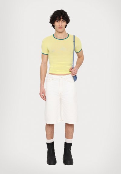 Top jaune ajusté avec des accents teal, shorts blancs jusqu'aux genoux et bottes noires. Le modèle porte un petit sac bleu. Design simple et minimaliste.