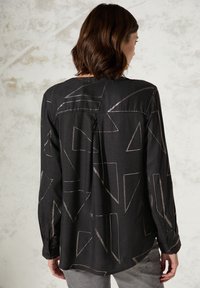 Chemise noire avec un motif triangulaire géométrique en fil clair. Manches longues, poignets à bouton, et coupe décontractée avec une fente au dos.