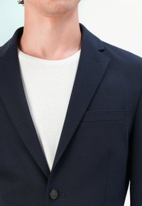 Blazer azul-marinho feito de tecido liso, com colarinho notched, fecho de um botão e um bolso no peito, usado sobre uma camisa branca.