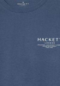 Plava pamučna majica s okruglim izrezom; sadrži bijeli tekst: "HACKETT LONDON" i dodatne detalje ispod. Glatka tekstura, ležeran dizajn.