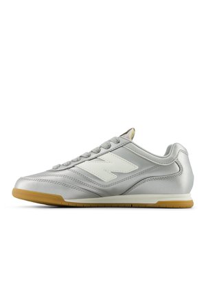 Sølv lavtop sneaker med hvidt logo på siden, grå snørebånd og en lysebrun gummisål, vist fra højre profil.