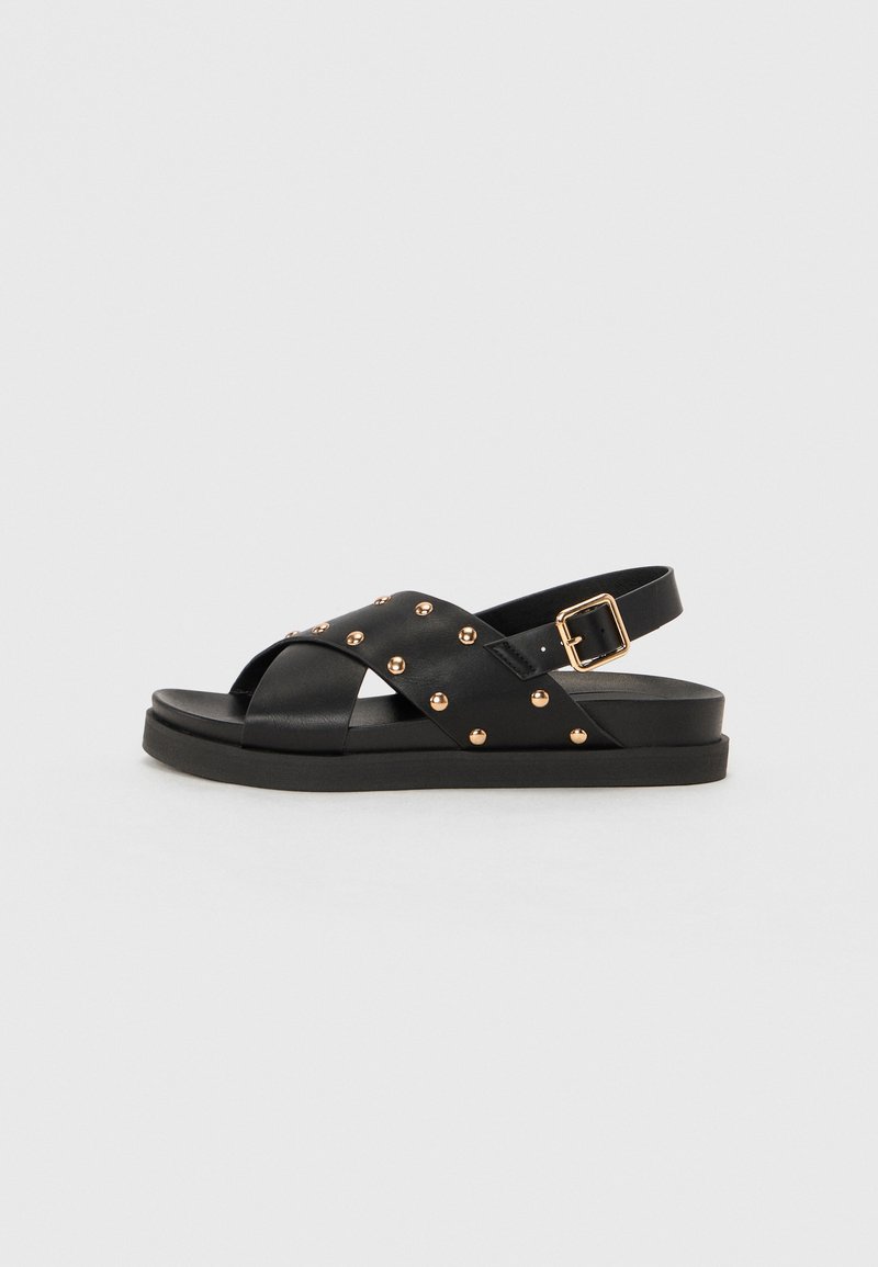 Black sandal with crisscross strap design, featuring gold studs and a buckled ankle strap; flat sole with a smooth texture.