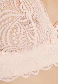 Reggiseno in pizzo di un delicato rosa con intricati motivi floreali, bordi ondulati e un delicato fiocco decorativo. Il tessuto testurizzato ne esalta l'appeal visivo.