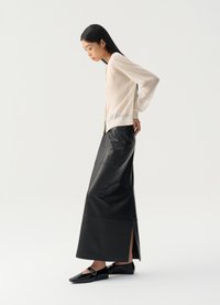 Blusa transparente bege clara, saia maxi de couro preta com abertura lateral e mocassins pretos brilhantes. A blusa tem um corte relaxado com mangas balão.