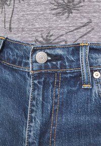 Vaqueros de denim con una cintura ajustada, tela azul, costuras naranjas y un botón de metal plateado en el centro, justo encima de la cremallera.