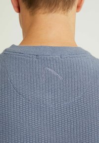 Bakvy av en person som bär en texturerad blå tröja med en liten broderad triangeldetalj på övre delen av ryggen.