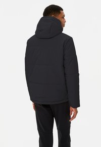 Zwarte pufferjacket met een capuchon, voorzien van een gewatteerd ontwerp, elastische manchetten en een rechte zoom. Draagt met zwarte broek.