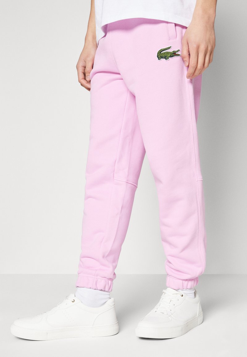 Pantalones de chándal rosa hechos de un tejido suave, con un logo de cocodrilo verde bordado en el muslo. Cuentan con puños elásticos y bolsillos laterales.