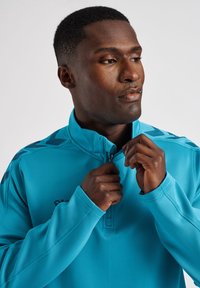 Maglietta sportiva da uomo con zip, di un brillante color turchese, realizzata in tessuto liscio con colletto alto e leggere decorazioni a motivi sulle spalle.