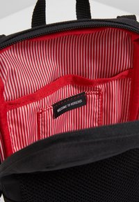 Intérieur de sac à dos noir avec doublure rayée rouge et blanche, comportant une petite poche étiquetée "WELCOME TO HERSCHEL".