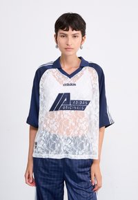 Balta džersija ar mežģīņu pārklājumu un tumši zilu apkakli un piedurknēm, ar lielu tumši zilu Adidas logotipu. Brīva forma ar sānu svītrām.