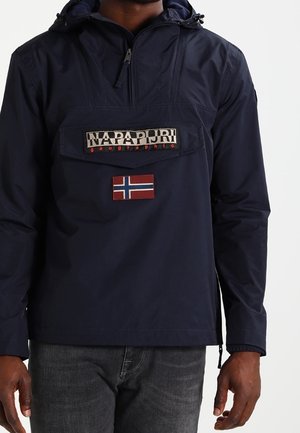 Marineblauer Napapijri Pullover mit Fronttasche, Reißverschluss und norwegischem Flaggenabzeichen, getragen zu dunklen Jeans.