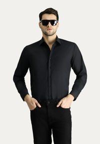 Camicia nera a maniche lunghe realizzata in tessuto liscio, con bottone frontale, colletto e una singola tasca sul petto, abbinata a pantaloni neri.