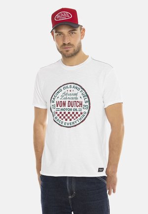 T-shirt en coton blanc avec un imprimé rond vieilli en rouge et vert, comprenant du texte et une bordure à damiers. Porté avec un jean en denim foncé.