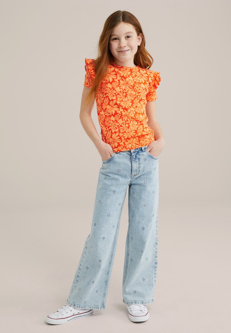 Spilgti oranžs ziedu motīvu īsā piedurkne krekls ar frillēm, saderēts ar gaiši ziliem platām kājām džinsiem ar izšūtiem ziedu elementiem. Baltas kedas.