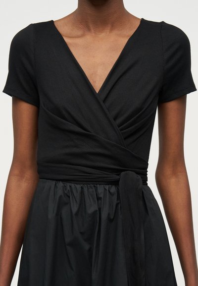 Γυναίκα που φοράει μαύρο κοντομάνικο μπλουζάκι με ντυμένο ντεκολτέ V και δεμένη ζώνη στη μέση, συνδυασμένο με φούστα με πιέτες.