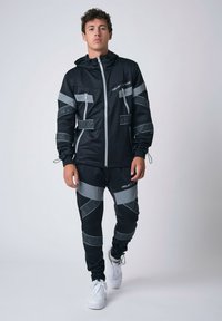 Conjunto deportivo negro que incluye una chaqueta con capucha y cremallera con detalles reflectantes y pantalones ajustados a juego, ambos con bolsillos.