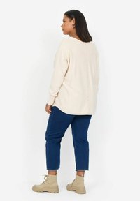 Beige gestrickter Oberteil mit V-Ausschnitt und Knopfdetail auf der Rückseite, kombiniert mit blauen, knielangen Jeans und beigen Stiefeletten.