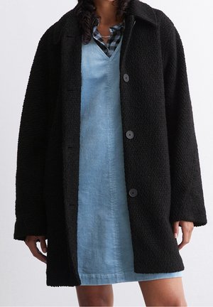 Femme portant un manteau noir texturé par-dessus une robe en velours côtelé bleue et une chemise à carreaux noire et bleue, avec un collier chaîne en or.