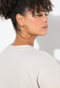 Beige Übergrößenhemd mit weicher Textur, kombiniert mit großen, metallisch goldenen Creolen. Schatten erzeugen Tiefe vor einem neutralen Hintergrund.