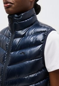 Κοντινή λήψη ενός ατόμου που φοράει λαμπερό, ναυτικό μπλε φουσκωτό γιλέκο με ραφές πάνω από λευκό κοντομάνικο πουκάμισο, με το γιλέκο φερμουάρ κλειστό.