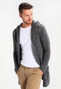 Cárdigan gris de punto con capucha, que presenta un patrón texturizado y un diseño de frente abierto; combinado con una camisa blanca y pantalones beige.