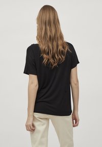 Kvinna med långt, vågigt ljusbrunt hår som bär en svart topp med korta ärmar och beige byxor, stående vänd bort från kameran mot en enfärgad bakgrund.