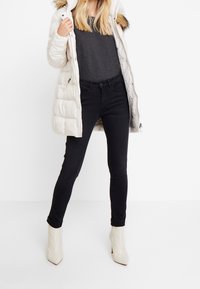 Biała puchowa kurtka z futrzanymi akcentami, szary dzianinowy top, czarne obcisłe jeansy oraz kremowe botki na wysokim obcasie. Dopasowana sylwetka i gładka faktura.