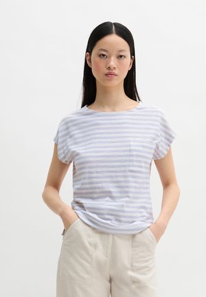 Jonge vrouw met steil zwart haar, gekleed in een lichtblauw-wit gestreept T-shirt met zakje en beige broek, handen in de zakken, neutrale achtergrond.