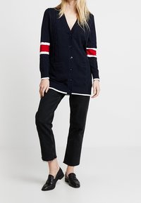 Marineblauwe gebreide cardigan met kabelpatroon, rode en witte strepen op de mouwen, knoopsluiting, voorkruiszakken en een ontspannen pasvorm, gecombineerd met zwarte jeans.