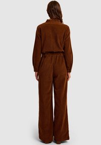 Brązowy kombinezon z sztruksu z luźnymi spodniami, długimi rękawami i elastycznym pasem. Posiada pionową, prążkowaną strukturę i guziki przy kołnierzyku.