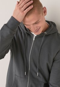 Sudadera con cremallera de color gris oscuro, con bolsillo tipo canguro, capucha con cordones y cremallera plateada. Presenta un tejido suave y texturizado.