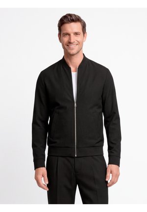 Lächelnder Mann, der eine schwarze Jacke mit Reißverschluss über einem weißen Hemd und schwarze Hosen trägt, vor einem schlichten weißen Hintergrund stehend.