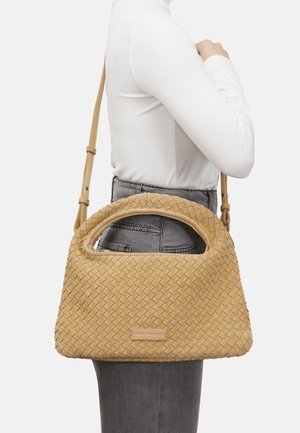 Frau trägt weißen Langarmpullover und graue Hose und hat eine große beigefarbene, geflochtene Handtasche mit kurzen und langen Trägern über der Schulter hängen.