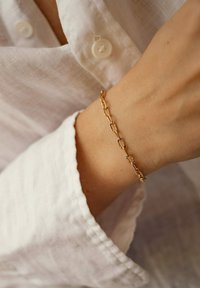 Pulsera de cadena de oro con eslabones ovalados y acentos texturizados, llevada en una muñeca, contrastando con una camisa de lino blanca.